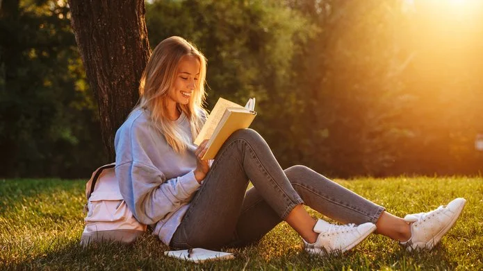 portrait of a delighted young woman sitting on a grass and reading a book
